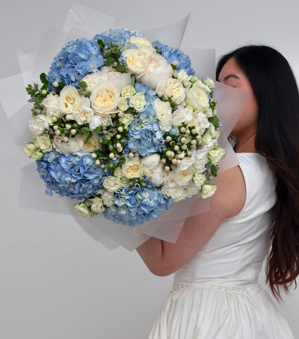 Photograph shows a woman in a white dress holding out 'Blue Light' bouquet. The bouquet features a mix of ivory and baby blue flowers.