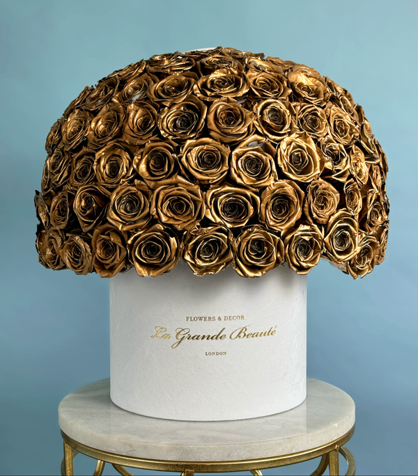 A dome shaped arrangement of preserved gold roses in a white 'La Grande Beauté' branded box. The arrangement is on top of a marble topped stool on a light blue background.