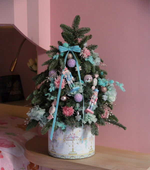 Decorative Christmas tree with pastel ornaments in a decorative box against a pink wall.