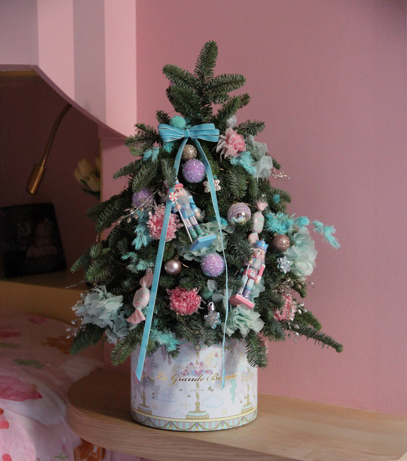 Decorative Christmas tree with pastel ornaments in a decorative box against a pink wall.