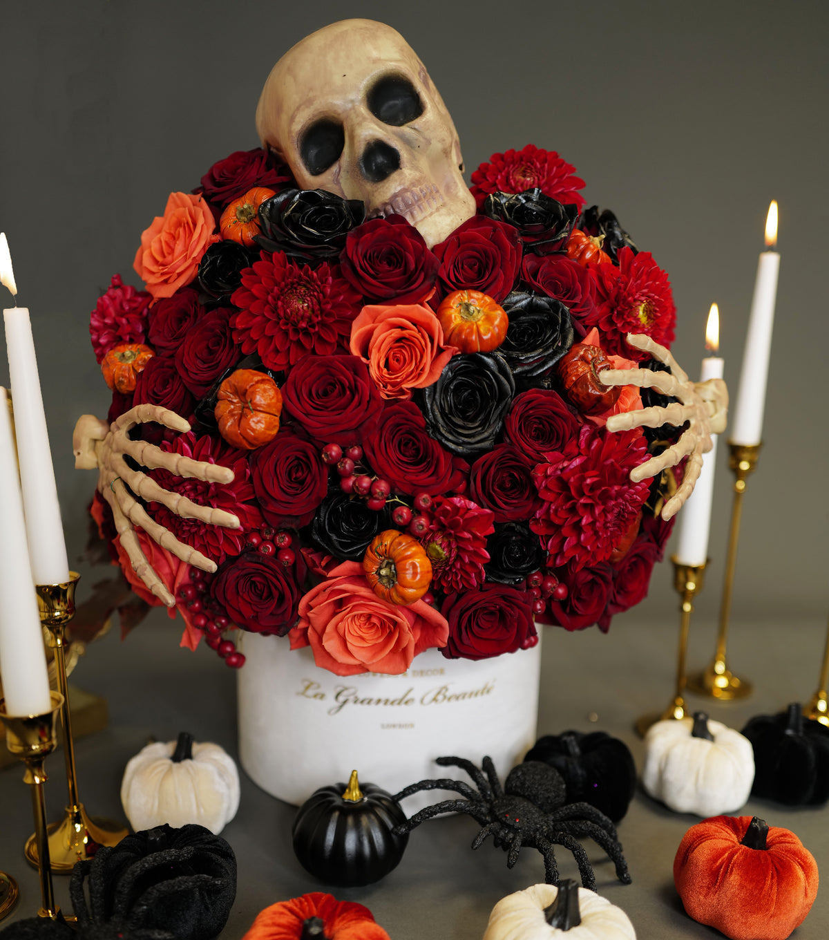 A decorative arrangement featuring a skeleton hand holding a bouquet of red and black flowers, with miniature pumpkins and black candles on a table.